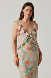 A woman is wearing a sleeveless, fitted dress with adjustable shoulder straps tied into bows. The dress features a colorful, abstract floral pattern in shades of pink, orange, yellow, green, and beige, with ruffled detailing along the neckline and shoulders.